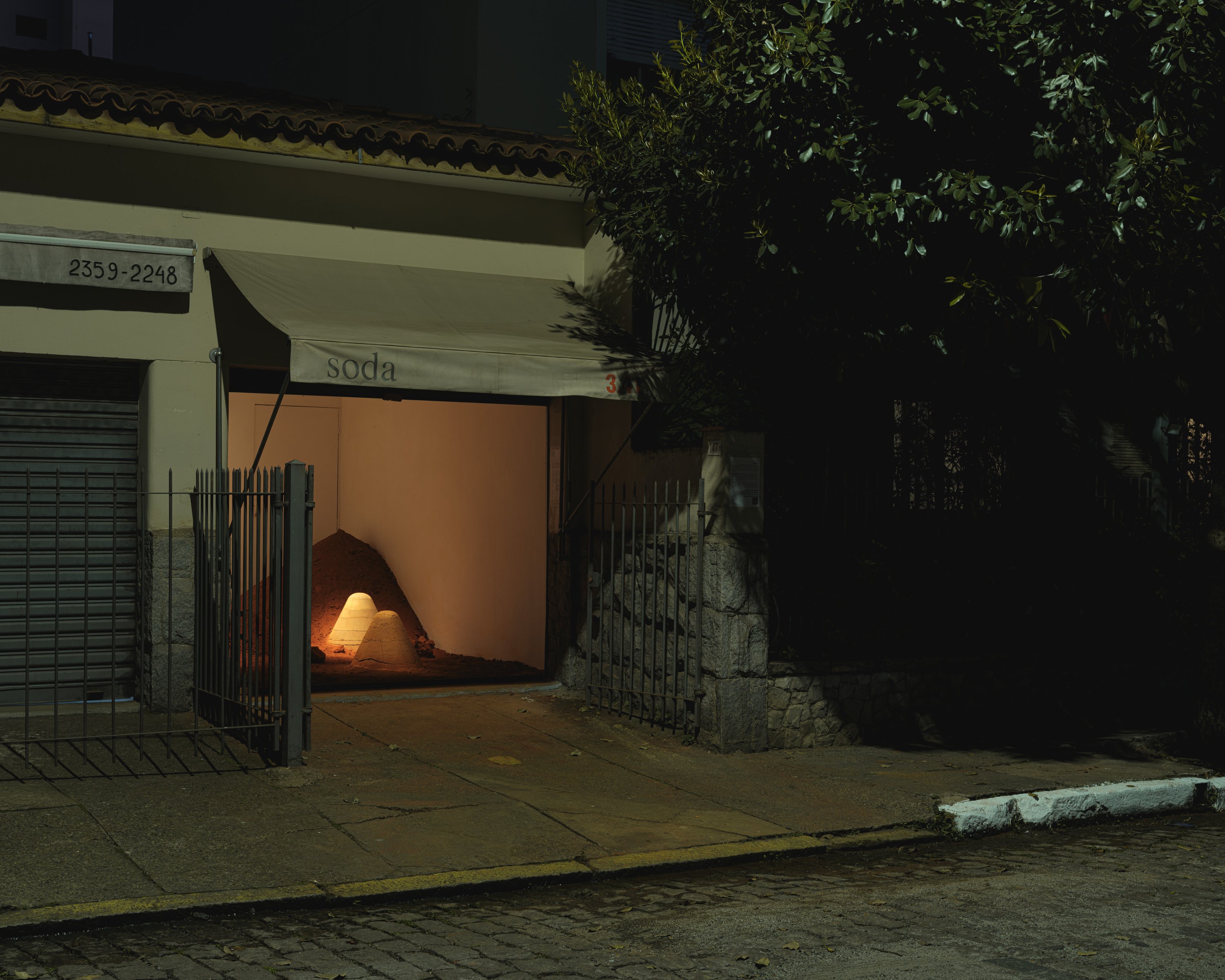 Night street under a soda awning, an open garage glowing onto a sand mound and two pale cones behind an iron gate — 3.4t installation in São Paulo by Arquipelago Arquitetos, curated by Duto at Espaço Soda (2026), a lit room on a dark sidewalk