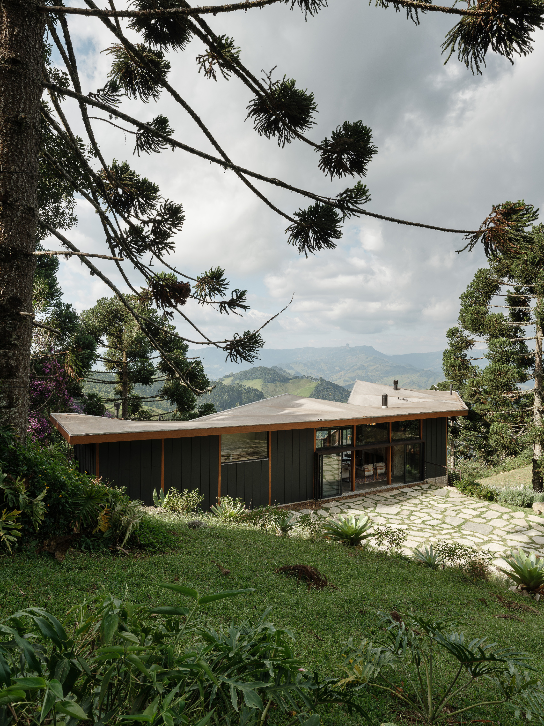 Araucaria branches frame a dark timber pavilion with a folded roof and a stone path, mountains beyond the lawn — Arundú Refuge in Sapucaí-Mirim by Estúdio HAA! with landscape by Olivia Uliano (2026), a highland house on the Mantiqueira slope