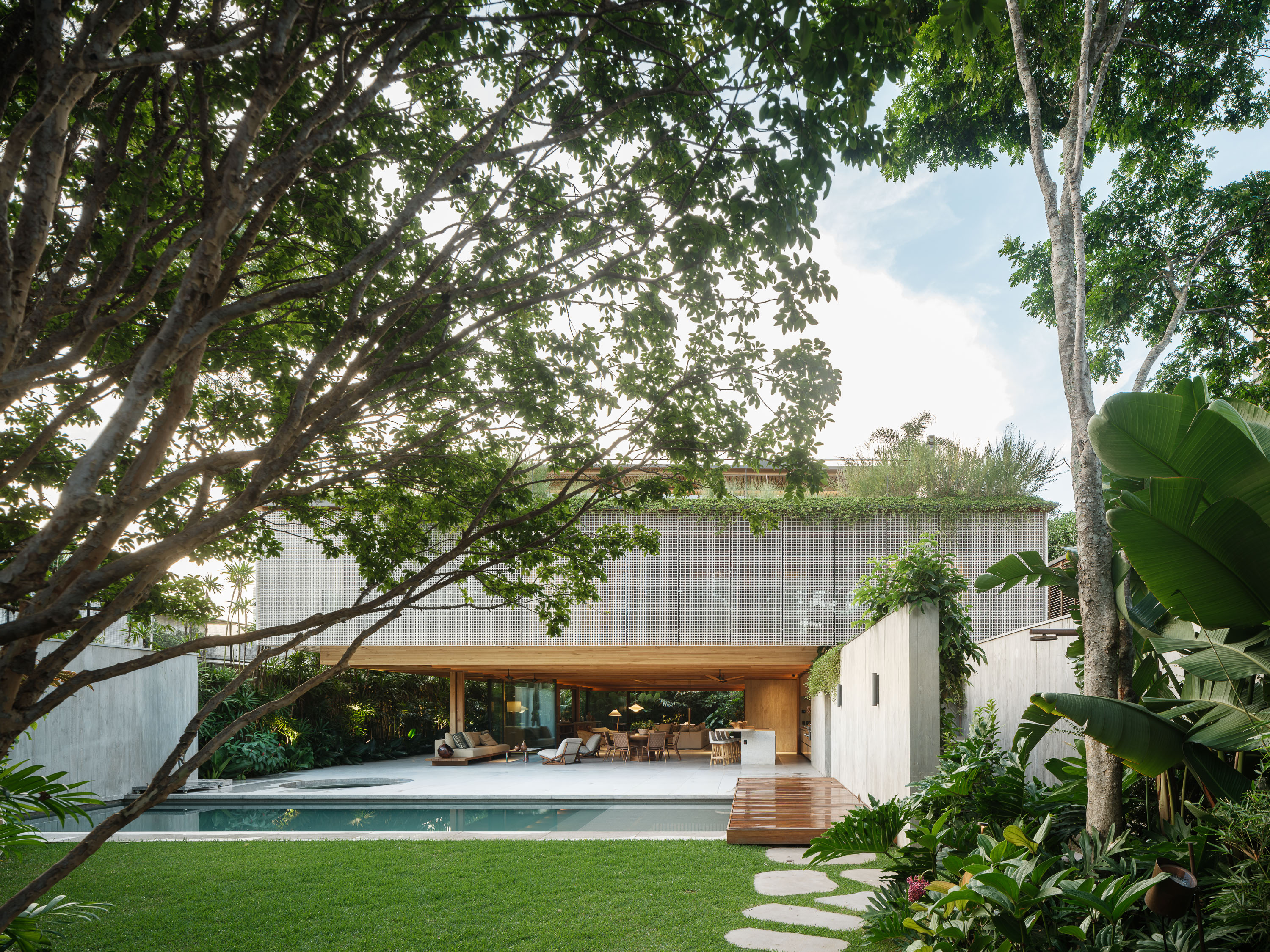 Pool and lawn framed by tree canopy toward a glazed living floor under a screened upper volume with a green roof — JGB House in São Paulo by Bernardes with landscape by Rodrigo Oliveira Paisagismo, a timber house opening fully to the garden